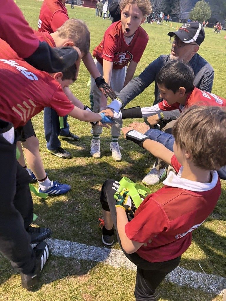 Chris kneeling in a hands-in huddle with his flag-football team in red 49ers jerseys